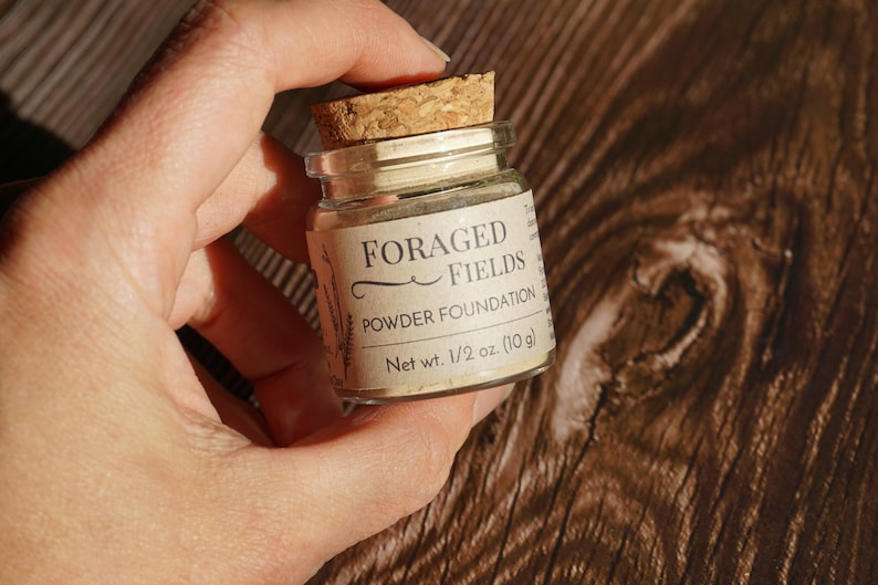 May include: A small glass jar with a cork stopper, labeled "FORAGED FIELDS Powder Foundation." The label indicates a net weight of 1/2 oz (10g). The jar is held in a hand against a wooden background.