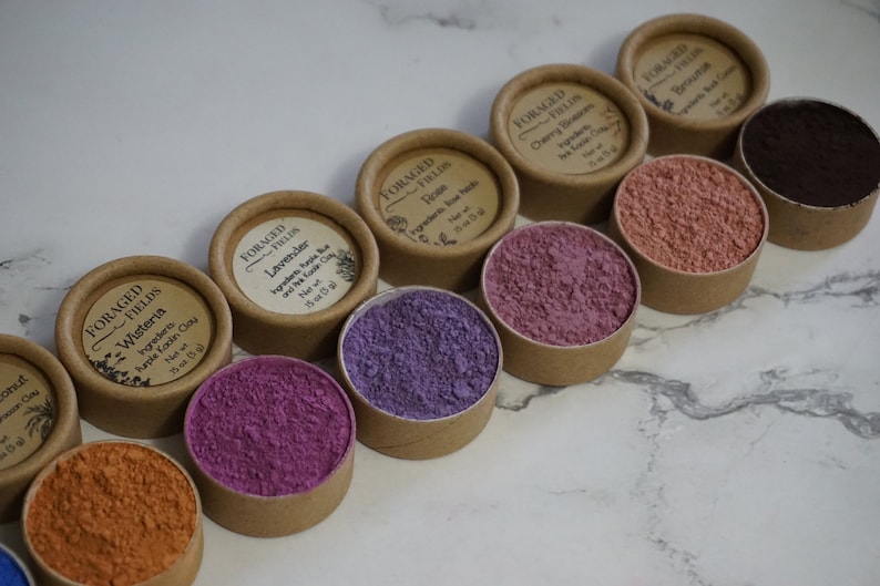 May include: A collection of small, round containers with lids, each filled with a different colored powder. The containers are labeled with the words "Foraged Fields" and the name of a flower or herb, such as "Wisteria", "Lavender", and "Rose".