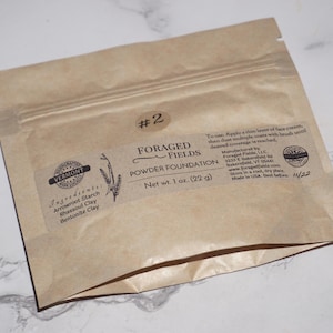 May include: A brown paper packet of Foraged Fields Powder Foundation, net weight 1 oz (22 g). The label includes the ingredients: arrowroot starch, rhassoul clay, and bentonite clay. The packet has a zip closure and the text "#2".