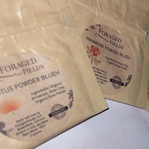 May include: Close-up of three paper packets with circular labels. The labels read "Foraged Fields" and list the product name: Lotus, Primrose, and Peach Powder Blush. Ingredients are listed. The packets are beige.