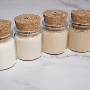 May include: Five small glass jars with cork stoppers, each containing a different shade of powder. The jars are labeled #0 through #4. The powders range in color from white to a light brown.