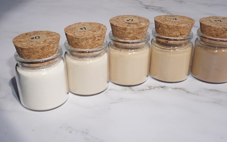 May include: Five small glass jars with cork stoppers, each containing a different shade of powder. The jars are labeled #0 through #4. The powders range in color from white to a light brown.