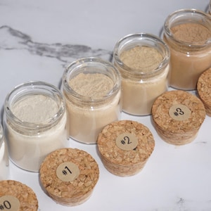 May include: A collection of small glass jars filled with various shades of powder, ranging from white to brown. Cork stoppers with numbered labels sit beside each jar. The jars are arranged on a white and gray marble surface.