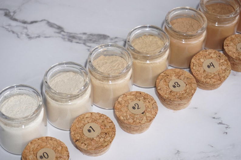 May include: A collection of small glass jars filled with various shades of powder, ranging from white to brown. Cork stoppers with numbered labels sit beside each jar. The jars are arranged on a white and gray marble surface.