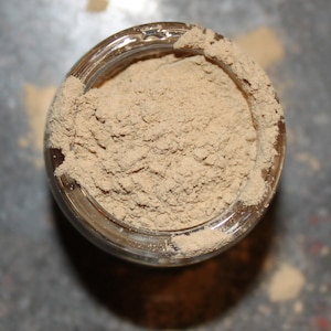 May include: Overhead view of a clear glass jar filled with a light brown powder. The powder is finely ground and fills the jar to the brim, with some spilling over the rim. The jar sits on a surface, casting a shadow.