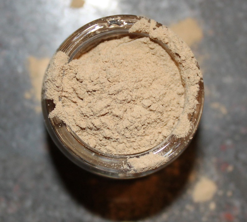 May include: Overhead view of a clear glass jar filled with a light brown powder. The powder is finely ground and fills the jar to the brim, with some spilling over the rim. The jar sits on a surface, casting a shadow.
