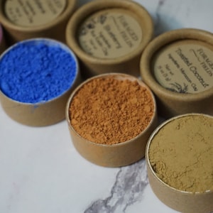 May include: Seven small round containers with lids, each containing a different colored powder. The powders are shades of purple, blue, orange, brown, and beige. The lids are labeled with the text "Foraged Fields" and the name of the powder.