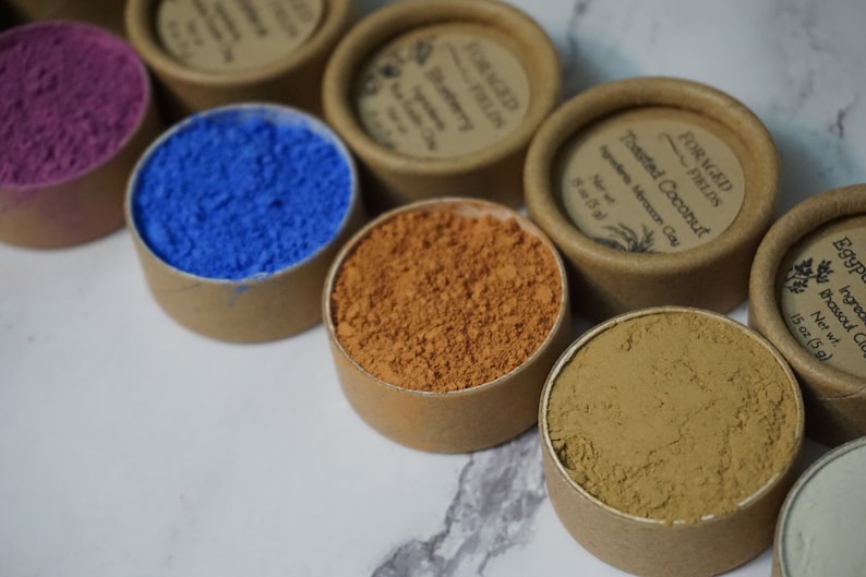 May include: Seven small round containers with lids, each containing a different colored powder. The powders are shades of purple, blue, orange, brown, and beige. The lids are labeled with the text "Foraged Fields" and the name of the powder.