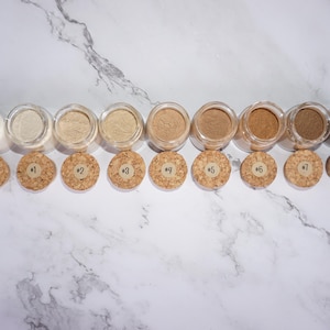 May include: A collection of nine small, clear glass jars filled with various shades of powder, ranging from light beige to dark brown. Each jar has a cork stopper with a number from 0 to 8 printed on it. The jars are arranged in a row on a white marble surface.