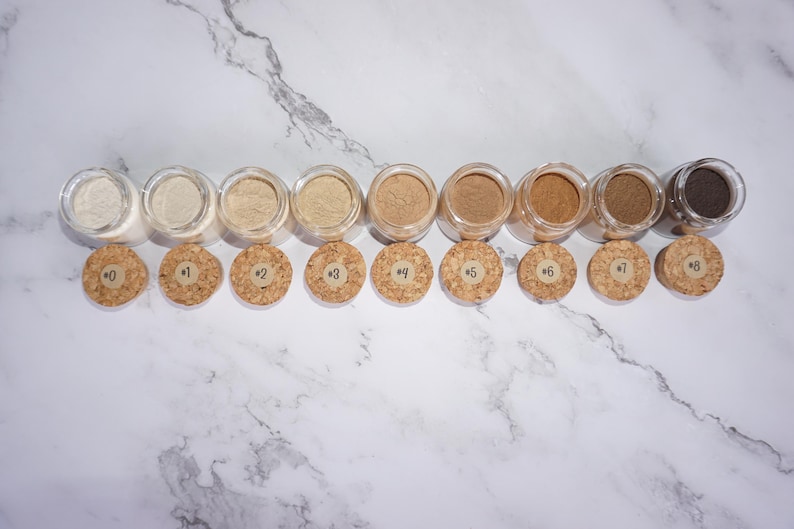 May include: A collection of nine small, clear glass jars filled with various shades of powder, ranging from light beige to dark brown. Each jar has a cork stopper with a number from 0 to 8 printed on it. The jars are arranged in a row on a white marble surface.