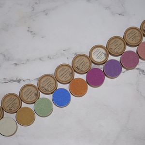 May include: A row of 14 small round containers with lids, each containing a different colored powder. The containers are made of brown cardboard and have a label with the text "FORAGED" and "ORGANIC COLOR".