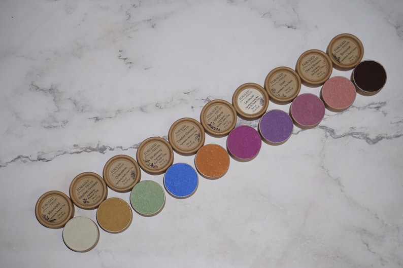 May include: A row of 14 small round containers with lids, each containing a different colored powder. The containers are made of brown cardboard and have a label with the text "FORAGED" and "ORGANIC COLOR".