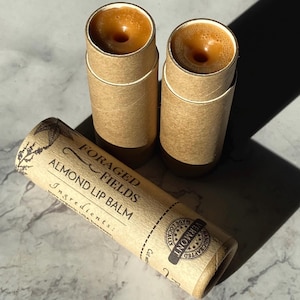 May include: Two brown cardboard tubes with a brown lip balm inside. The tubes are labeled "Foraged Fields Almond Lip Balm" and have a brown stamp that says "Handcrafted Locally" with a star design.