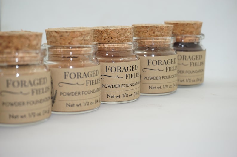 May include: A collection of small glass jars with cork stoppers, filled with powder foundation. Each jar has a label that reads "FORAGED FIELDS POWDER FOUNDATION" and "Net wt. 1/2 oz. (14g)". The jars are arranged in a row against a white background.