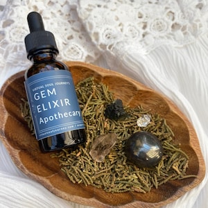 Könnte beinhalten: Eine kleine braune Glasflasche mit Tropfer, beschriftet mit "Gem Elixir Apothecary" und "Virtual Soul Journey's". Die Flasche steht in einer Holzschale mit grünen Kiefernnadeln, klaren Quarzkristallen und schwarzen Steinen.