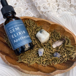 May include: A dark glass bottle with a dropper top, labeled "Gem Elixir Apothecary" and "Virtual Soul Journey's". The bottle is sitting in a wooden bowl filled with green cedar shavings and various crystals.