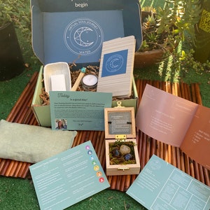 Puede incluir: Una caja azul verdosa con las palabras "Virtual Soul Journey" y "Water" en la tapa. La caja contiene una variedad de artículos, incluyendo un pequeño tazón blanco, una vela, un palo de madera, un cuaderno, una pequeña caja de madera con cristales y algunos papeles con texto. La caja está sentada sobre una estera tejida marrón.