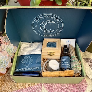 Pode incluir: Uma caixa verde e azul com as palavras "Virtual Soul Journey" na tampa. A caixa contém uma variedade de itens, incluindo uma vela, uma pequena caixa de madeira, uma garrafa de óleo essencial, um bastão de sálvia e uma pequena bolsa de cristais. A caixa está sentada em uma superfície com padrão rosa e branco.