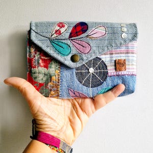 Könnte beinhalten: Eine Patchwork-Geldbörse mit Jeans-Basis und farbenfrohen Stoffakzenten. Der Umschlag hat gestickte Blattmotive in Rot, Rosa und Türkis. Ein brauner Knopf und ein Lederetikett sorgen für Details. Die Geldbörse wird in einer Hand gehalten.