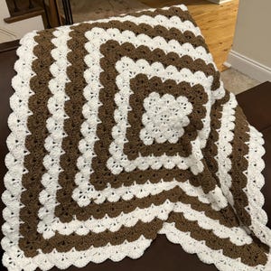 May include: A crocheted blanket with a brown and white pattern. The blanket is made with a scalloped edge and has a square design in the center.