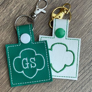 May include: Two green and white leather keychains with a Girl Scout logo. One keychain has the letters "GS" embroidered in white on a green background. The other keychain has a white outline of the Girl Scout logo on a white background. Both keychains have a silver and gold metal ring.