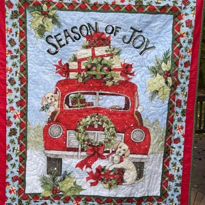 May include: A red vintage car with a wreath on the front bumper is filled with wrapped presents and driven by two dogs. The car is on a snowy background with a blue sky. The text "Season of Joy" is written in a decorative font above the car.