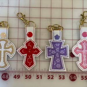 May include: Four keychains with different colored crosses. The crosses are embroidered with different patterns. The keychains are white with gold rings.