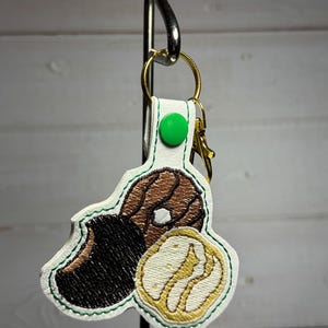 May include: A white keychain with a green button and gold hardware. The keychain features an embroidered design of a dachshund dog with brown and black fur and a cookie. The design is outlined in green.