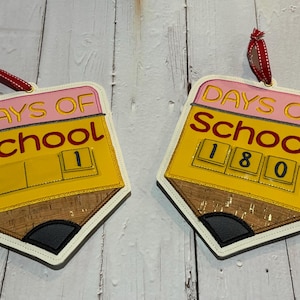 May include: Two yellow pencil-shaped ornaments with pink and red accents. The ornaments are decorated with the text "Days of School" and a number counter. One ornament shows the number 1, and the other shows the number 180.
