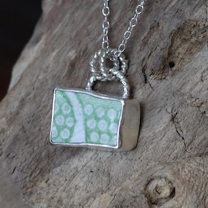 May include: A silver necklace featuring a rectangular pendant with a light green and white patterned design. The pendant is encased in a silver frame and hangs from a delicate silver chain. The necklace is displayed on a piece of weathered wood.