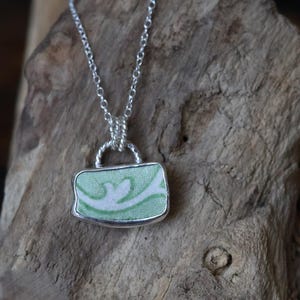 May include: A silver necklace with a rectangular pendant. The pendant features a light green and white patterned design, encased in a silver frame. The necklace is displayed against a textured, wooden background.