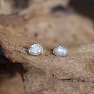 Può includere: Un paio di orecchini a bottone con perle. Ogni orecchino presenta una perla bianca di forma irregolare incastonata in una lunetta d'argento. Gli orecchini sono esposti su una superficie di legno marrone strutturata.