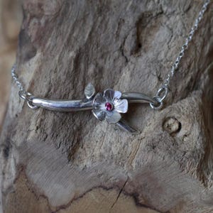 May include: A silver necklace featuring a floral design. The pendant is a stylized branch with a flower and a small pink gemstone in the center. The necklace is on a silver chain.