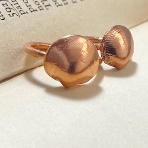 Op de afbeelding: Twee koperen ringen met een schelpmotief. De ringen zijn gemaakt van een glanzend, roodbruin metaal. Eén ring heeft een gladde, afgeronde schelpvorm, terwijl de andere een getextureerde, geribbelde schelpvorm heeft. De ringen zijn geplaatst tegen de achtergrond van een boek.