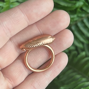 May include: A gold-colored ring with a detailed, textured design. The ring features a rounded band and a decorative top with a ridged, oblong shape. The ring is held in a hand against a blurred green background.