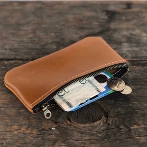 May include: A brown leather zippered pouch with a zipper pull that says "YKK". The pouch is open and contains cash, a credit card, and coins.