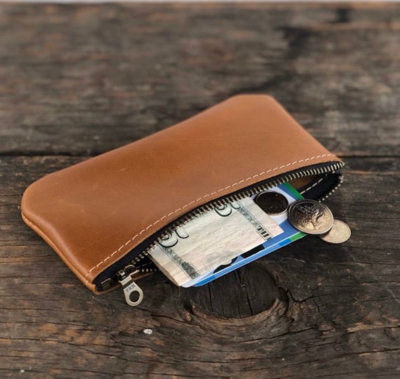 May include: A brown leather zippered pouch with a zipper pull that says "YKK". The pouch is open and contains cash, a credit card, and coins.
