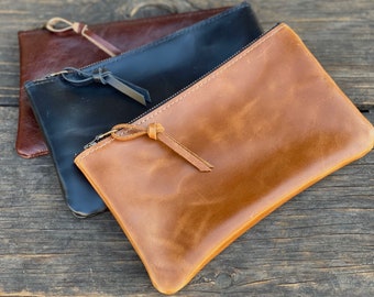 May include: Three leather zipper pouches in different colors: brown, black, and dark brown. The pouches are stacked on top of each other, with the brown pouch on top.