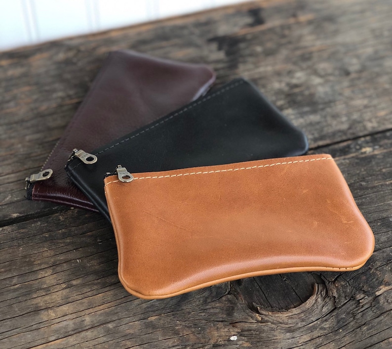 May include: Three leather zipper pouches in brown, black, and tan. The pouches are stacked on top of each other, with the tan pouch on top.