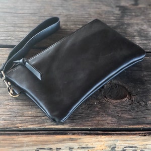 以下が含まれることがあります： 手首ストラップとジッパー留め具が付いた黒い革製のクラッチバッグ