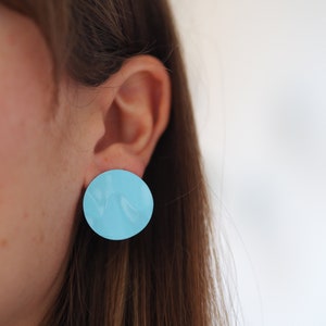 Puede incluir: Un pendiente grande y redondo de color azul claro se muestra en la oreja de una persona. El pendiente tiene un acabado brillante y una superficie ligeramente texturizada. Diseño sencillo y moderno.