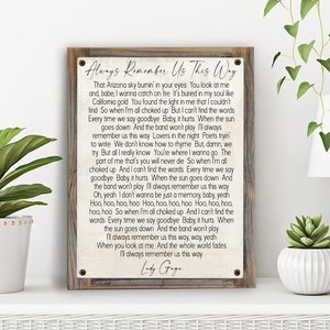 May include: A rustic wooden sign with a white background and brown wood frame. The sign features the lyrics to the song "Always Remember Us This Way" by Lady Gaga. The lyrics are printed in black text and include the lines "That Arizona sky burnin' in your eyes" and "I'll always remember us this way."