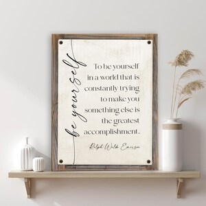 May include: A framed wall art piece with the quote "To be yourself in a world that is constantly trying to make you something else is the greatest accomplishment." The artwork is on a wooden shelf with decorative items.