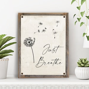 May include: A rustic wooden sign with a white background and black text that reads "Just Breathe". A black and white dandelion with seeds blowing in the wind is depicted above the text.