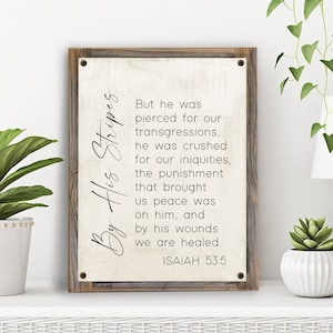May include: A rustic wooden sign with a white background and black text. The text reads "By His Stripes" and a Bible verse from Isaiah 53:5. The sign is a religious decor item.