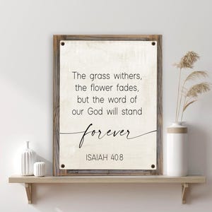 May include: A framed wooden sign with the text "The grass withers, the flower fades, but the word of our God will stand forever" and the reference "ISAIAH 40:8". The sign is displayed on a wooden shelf with decorative items.