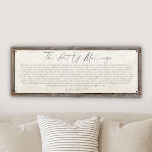 May include: A rectangular wooden sign with the text "The Art Of Marriage" in elegant script. The sign features a cream-colored background with printed text and is framed by a rustic, weathered wooden border. The sign is mounted on a white wall.