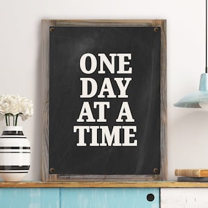 May include: A black chalkboard sign with a wooden frame, featuring the text "ONE DAY AT A TIME" in white lettering.