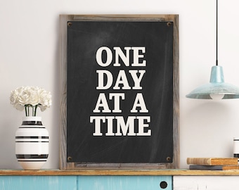 May include: A black chalkboard sign with a wooden frame, featuring the text "ONE DAY AT A TIME" in white lettering.