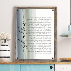 May include: A framed print with a watercolor background and lyrics from the song "Shallow" by Lady Gaga and Bradley Cooper. The lyrics are printed in a white font and the word "shallow" is written in a large, black font on the left side of the print.